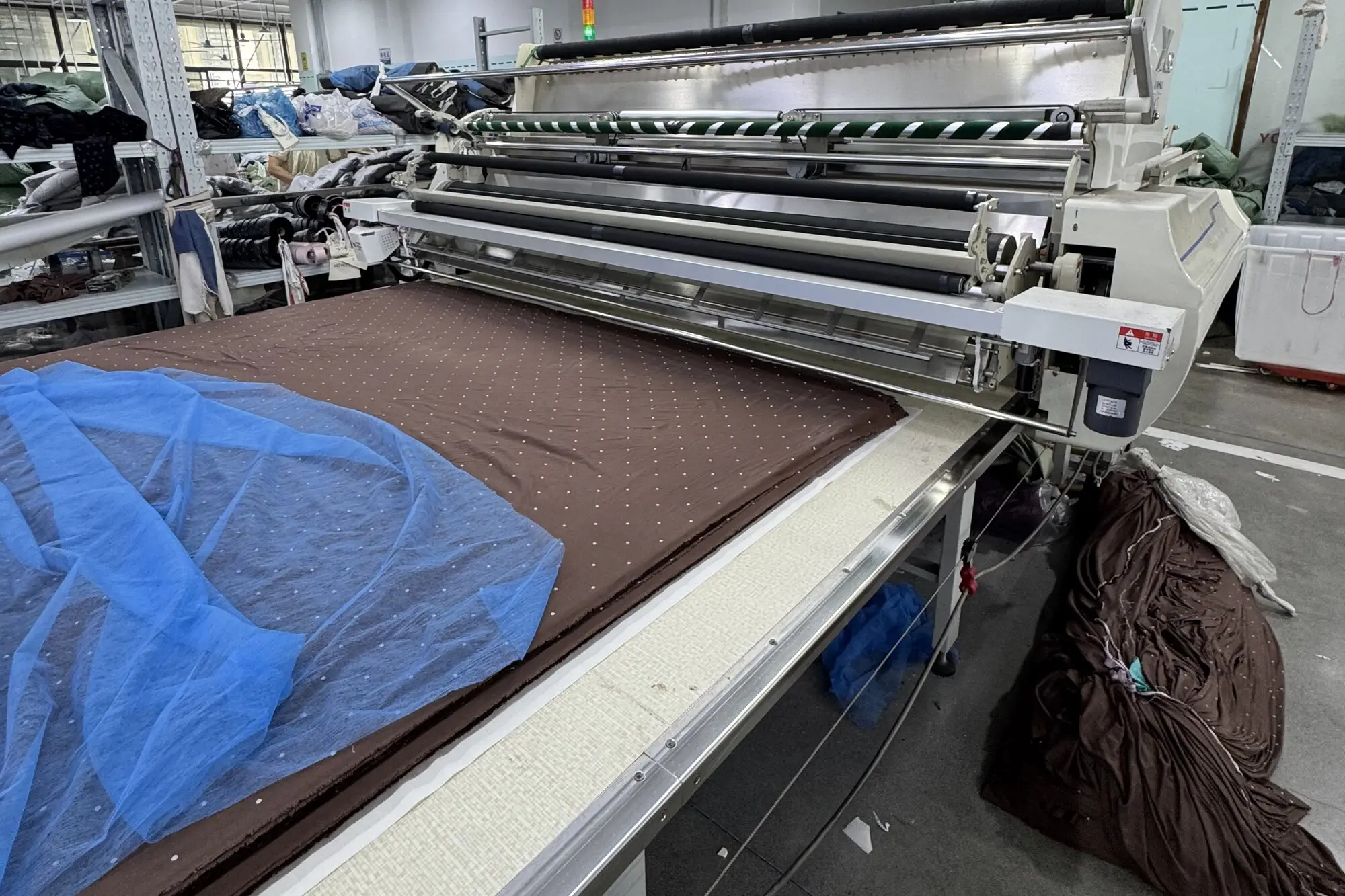 automatic fabric spreading machine laying a brown dotted fabric ply along a long cutting table on the ApronCraft factory floor