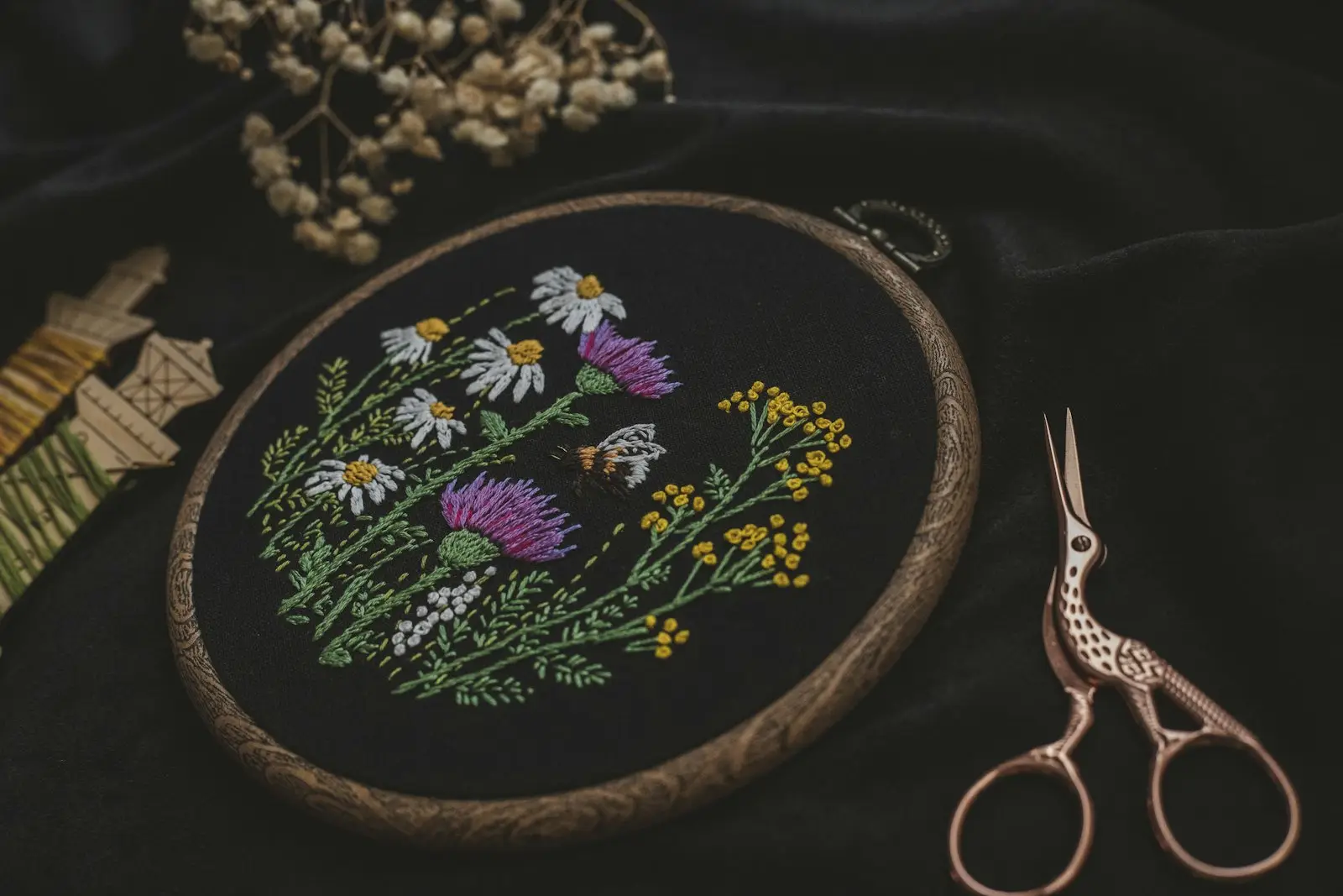 floral embroidery stitched in a wooden hoop on black fabric, with stork scissors and thread spools beside it