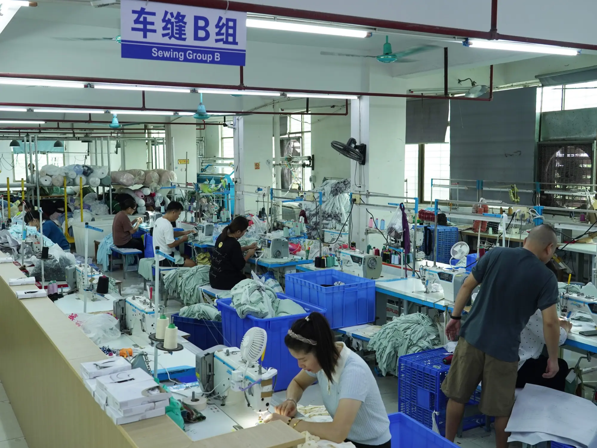 operators at rows of industrial sewing machines in the ApronCraft Sewing Group B workshop, bundles of cut panels in blue bins beside each station