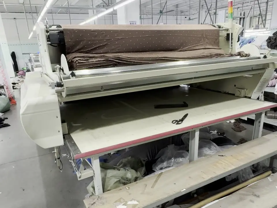 automatic fabric spreading machine laying a brown fabric ply along a long cutting table on the ApronCraft factory floor